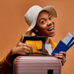 A woman holding a suitcase and plane tickets (Photo by IC Production via Shutterstock)
